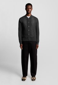 Grijze cardigan met een gestructureerd patroon, knoopsluiting aan de voorkant en kraag, gecombineerd met zwarte broek en gemusterde sneakers.