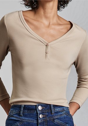 Personne portant un haut beige à manches longues boutonné et un jean bleu taille haute avec deux boutons métalliques visibles, mains posées sur les hanches.