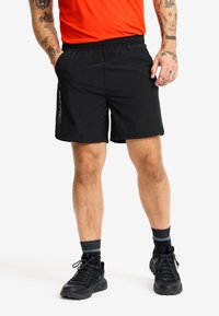 Svarta sportshorts med elastisk midja, utrustade med sidofickor och en reflektiv logodetalj. Bärs med svarta skor och strumpor.