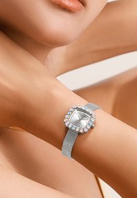 Montre-bracelet en maille argentée avec un cadran carré, ornée de gemmes incolores et pastel, dotée d'un cadran minimaliste et d'un bracelet texturé.