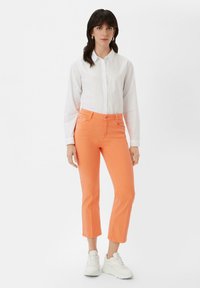 Pomarańczowe spodnie ściągane o dopasowanym kroju, z pięcioma kieszeniami i zapięciem na guzik, zestawione z białą koszulą zapinaną na guziki i białymi trampkami.
