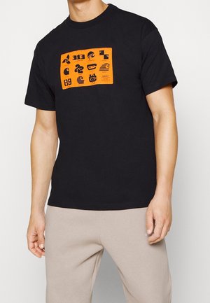 Persona que lleva una camiseta negra de manga corta con un gráfico rectangular naranja de varios logos negros y pantalones beige.