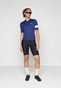 Marineblaue Radtrikot mit kurzen Ärmeln, Reißverschluss vorne, weißer Akzent am Ärmel. Schwarze Radhose und passende schwarze Schuhe. Sonnenbrille runden den Look ab.