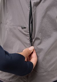 Leichte, graue Jacke mit strukturiertem Finish. Verfügt über einen schwarzen Reißverschluss und verstellbare Kordeldetails. Nahaufnahme einer Hand, die den Reißverschluss greift.