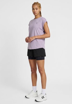 Kvinde med flettet hår iført en lys lilla ærmeløs skjorte, sorte shorts med inderleggings, hvide sokker og hvide sneakers, stående på hvid baggrund.