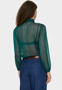 Blouse en tulle teal avec des manches bouffantes et un col à volants. Portée avec un pantalon en denim taille haute et une ceinture texturée marron.