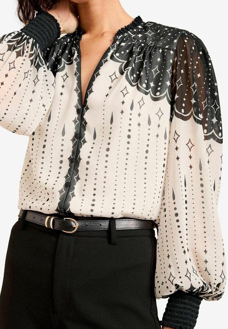 Blus i krämfärg med svart abstrakt mönster, med volangkrage och puffärmar. Svart texturerat bälte kompletterar skräddarsydda byxor.