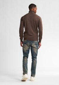 Pull en tricot marron avec un col montant, associé à un jean bleu délavé et des baskets blanches. Couture visible et deux poches arrière sur le jean.