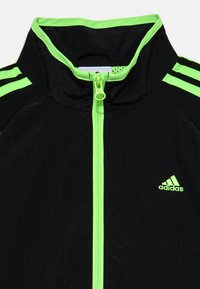 Crna jakna s visokim ovratnikom, zelenim detaljima i Adidas logotipom. Ima prednji zatvarač i teksturiranu tkaninu s horizontalnim linijama.
