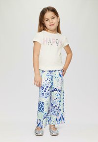 Lichtgekleurde t-shirt met "HAPPY" gedrukt in pailletten; patroon, wijde broek in blauwe en groene bloemenprint; zilveren platte schoenen.