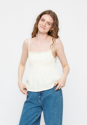 Νεαρή γυναίκα με κυματιστά μαλλιά, που φοράει λευκό αμάνικο τοπ με δαντέλα και φαρδιά μπλε τζιν, ποζάρει μπροστά σε απλό φόντο.