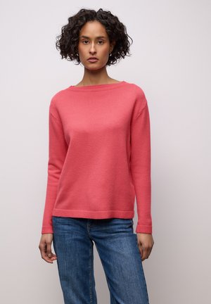 Frau mit lockigem, dunklem Haar, die einen rosa Pullover mit langen Ärmeln und blaue Jeans trägt, steht vor einem schlichten hellen Hintergrund.
