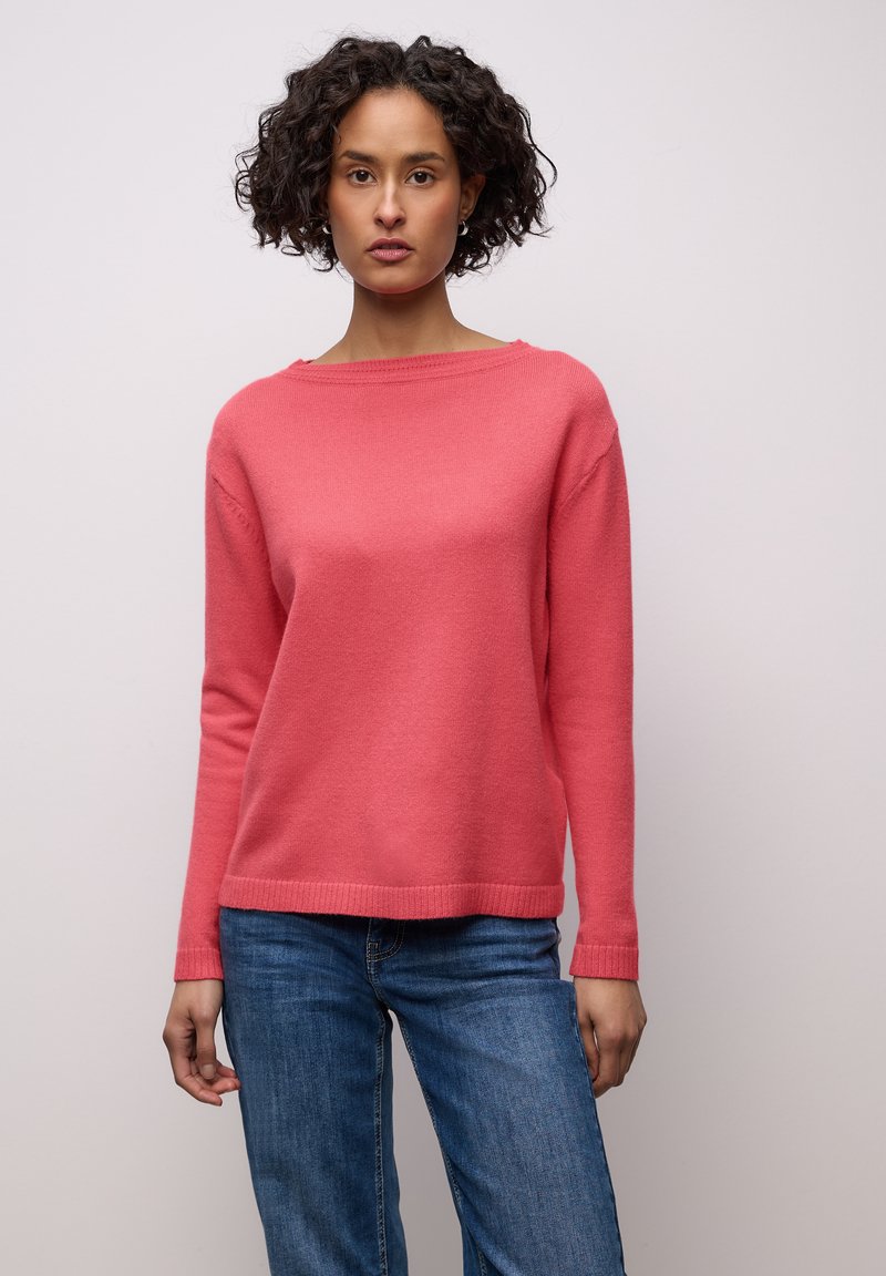 Femme aux cheveux bouclés et foncés, portant un pull rose à manches longues et un jean bleu, debout devant un fond clair uni.