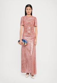 Lange roze paillettenjurk met korte pofmouwen en een nauwsluitende silhouet, gecombineerd met een blauwe en gouden clutch en crèmekleurige schoenen.