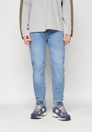 Jeans de mezclilla azul claro con un corte ajustado, combinados con una camiseta de manga larga gris que presenta detalles en verde oliva y zapatillas de running grises.