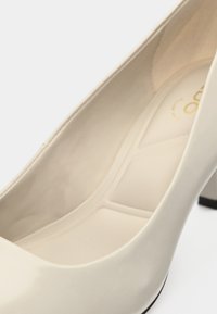 Pantof cu toc înalt din piele lacuită crem, cu vârf rotund și toc gros. Interiorul are un finisaj neted cu un logo subtil în auriu.