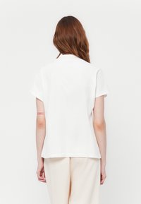 T-shirt bianco in cotone con maniche corte e taglio dritto. Presenta una parte posteriore texturizzata e un piccolo dettaglio testuale rosso sul braccio sinistro.