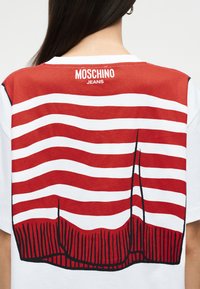 Πίσω όψη ατόμου που φοράει άσπρο μπλουζάκι με κόκκινες και λευκές κυματιστές ρίγες και μαύρες κάθετες γραμμές, με την επιγραφή "MOSCHINO JEANS" στο κέντρο πάνω μέρος.