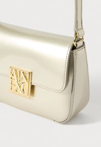 Torba iz kovinskega zlata z gladkim zaključkom, ki ima strukturirano obliko, preklopni pokrov in poudarjeno zlato logotipsko oznako na sprednji strani.