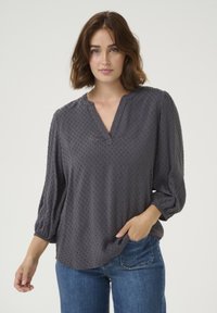 Grijze, gestructureerde blouse met een V-hals en pofmouwen, voorzien van een stippenpatroon. Gepaard met blauwe jeans, met de handen op de heupen.