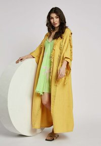 Lang, franjes kimono in geel gedragen over een lichtgroene jurk met roze bladpatronen, gecombineerd met bruine slidesandalen. Soepele textuur.
