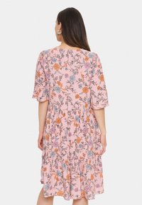 Rosa blommig klänning med trekvartslånga ärmar, lager-på-lager volangkant och mönster av orange och blå blommor. Mjuk tygtextur.