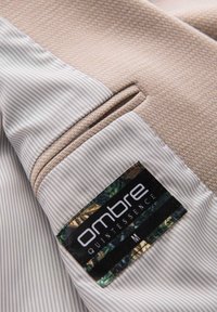 Veste beige texturée avec une doublure intérieure rayée et une étiquette noire cousue à l'intérieur qui porte la mention "ombre Quintessence M".