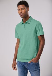 Camisa polo verde menta con una textura sutil, tapeta de tres botones y un pequeño logo en el pecho. Combinada con jeans azules.