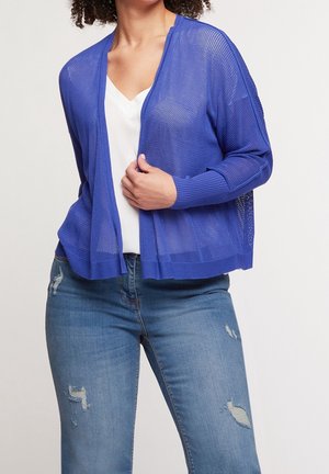 Femme portant un cardigan bleu en maille ajourée sur un top blanc et un jean bleu déchiré, vue du cou aux hanches sur un fond uni.