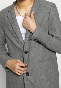 Grijze blazer met een gestructureerde stof, single-breasted ontwerp, met inkeping bij de revers, twee voorzakken en zwarte knoopaccenten.