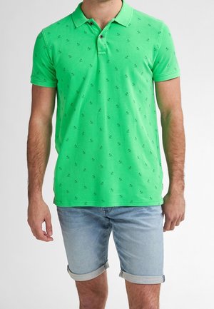 Mand iført en lys grøn kortærmet polo t-shirt med små mørke mønstre og lyseblå opbukte denimshorts.