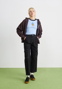 Pantaloni cargo neri con cuciture bianche, abbinati a una camicia azzurra chiara con una grafica colorata di fiamme e rose, e un felpa scura.