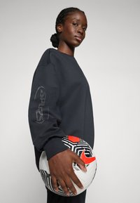 Felpa nera con polsini a costina, con un logo sul maniche. Il modello tiene un pallone da calcio a scacchi bianco e nero con accenti rossi.