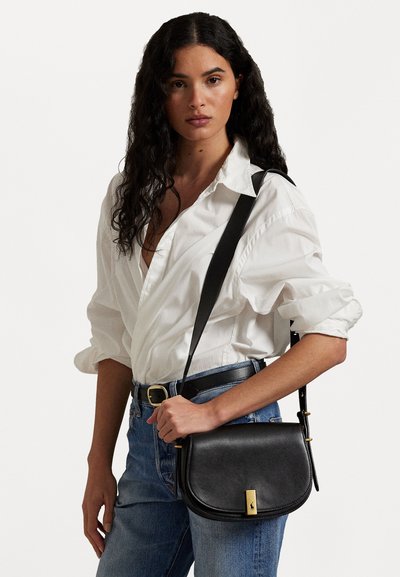 Sac en cuir noir porté en bandoulière avec une silhouette courbée, des accents en métal doré et une sangle ajustable, associé à une chemise blanche et un jean bleu.