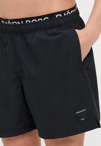 Calções de banho pretos feitos de tecido leve, com um cós elástico com logótipo, bolsos laterais e uma pequena etiqueta da marca na bainha.