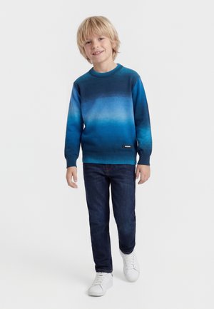 Garçon blond souriant, portant un pull bleu ombré, un jean foncé et des baskets blanches, debout devant un fond clair uni.