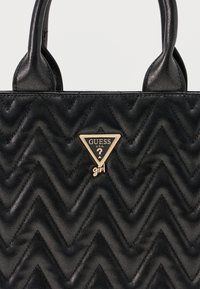 Μαύρη ραμμένη δερμάτινη τσάντα με μοτίβο σε σχήμα κεφαλαίων V, διπλές λαβές και επίχρυση πλακέτα με το λογότυπο Guess Girl στο κέντρο του εμπροστινού μέρους.