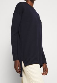 Marineblauer Strickpullover mit Rundhals, gerippten Säumen, überschnittenen Schultern und seitlichen Schlitzen, kombiniert mit einem strukturierten cremefarbenen Rock.