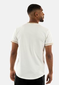 Camiseta blanca de manga corta y cuello redondo, con una marca "Project X Paris" acentuada en dorado en el cuello y los bordes de las mangas.