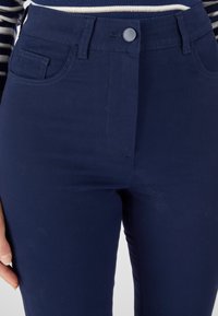 Marineblauwe broek van gladde stof, met een voorzak en een knoopsluiting, voorzien van zichtbare stikseldetails.