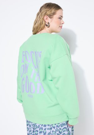 Mintgroene sweatshirt met een oversized fit, ronde hals en grote paarse tekst op de achterkant met de tekst "FOCUS ON THE GOOD PERIOD."