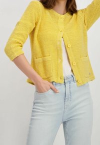 Gul stickad kofta med rund halsringning, trekvartslånga ärmar, framfickor och knappstängning. Buren över ljusblå jeans.