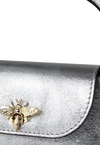 Sac à main argenté métallique avec un corps noir texturé et un emblème d'abeille doré sur le rabat, présentant des bords incurvés et des détails de couture.