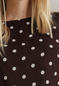 Ruskea kangas, jossa suuria valkoisia pilkkuja, suorakaiteen muotoinen pääntie ja lyhyet hihat, esittäen sileän tekstuurin.