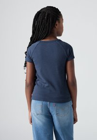 Meisje met lange vlechten, gekleed in een marineblauwe top met korte mouwen en lichtblauwe jeans, staand met haar rug naar de camera tegen een effen achtergrond.