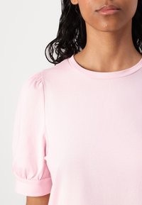 Femme aux cheveux bouclés foncés portant un t-shirt rose clair à manches courtes bouffantes et col rond.