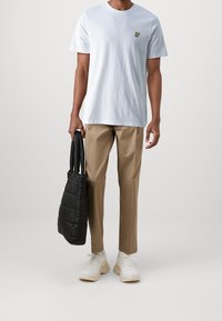 Helles, blaues Baumwoll-T-Shirt mit einem kleinen grünen Schmetterlingslogo, kombiniert mit beigen Hosen und einer schwarzen, gesteppte Tragetasche. Weiße Turnschuhe runden den Look ab.