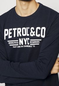 Muž v tmavě modré košili s dlouhým rukávem, na které je bílý nápis "PETROL & CO NYC Brooklyn U.S. Bez překážek Rychle a v čele od '73"