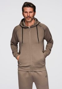 Homme barbu portant un sweat à capuche taupe zippé avec des cordons noirs et un pantalon de survêtement assorti, devant un fond uni.