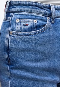 Κοντινό πλάνο μπλε τζιν με μπροστινή τσέπη που δείχνει ένα μικρό ορθογώνιο λογότυπο σε κόκκινο, λευκό και μπλε χρώμα και λεπτομέρειες με καρφίτσες.